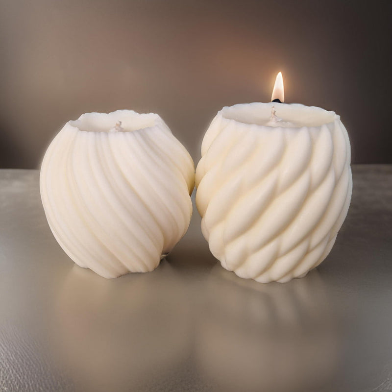 A white scented pillar candle with a twisted design, burning on a metal holder with a dark background.