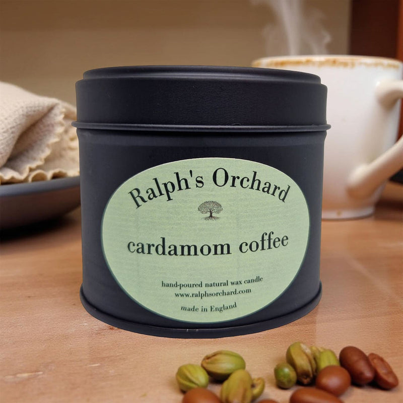 Candle tin labeled 'Ralph's Orchard cardamom coffee' on a wooden surface with coffee beans and a mug in the background.