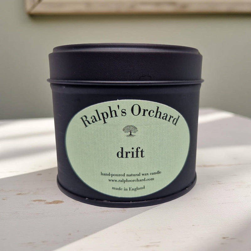 Candle in a black tin with 'Ralph's Orchard' label on a light wooden surface.