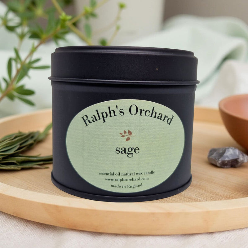 Candle in a black tin labeled 'Ralph's Orchard Sage' on a wooden surface with plants in the background.