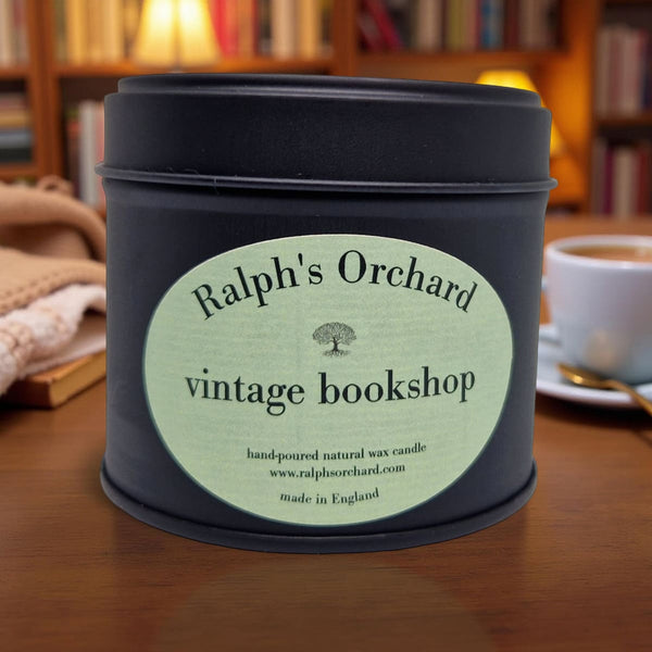 Candle in a tin labeled 'Ralph's Orchard vintage bookshop' on a wooden surface with a blurred background.