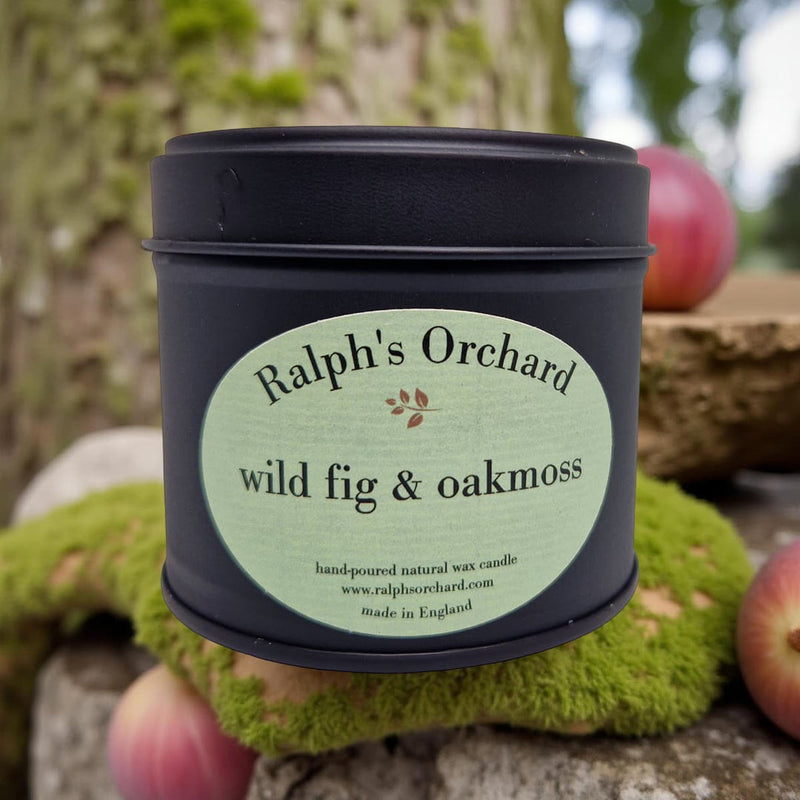 Candle in a black tin labeled 'Ralph's Orchard wild fig & oakmoss' against a natural background.