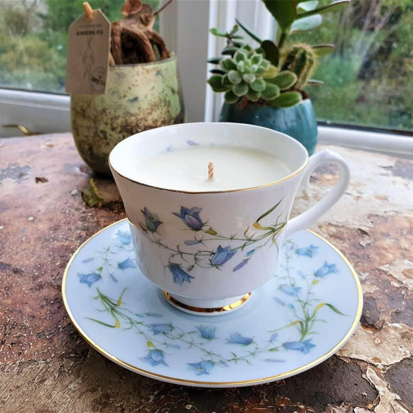Teacup and saucer with floral design containing a candle, placed on a wooden surface with plants in the background.