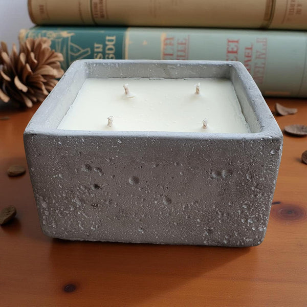 Square concrete candle with 4 wicks on a wooden surface with books and pine cones in the background.
