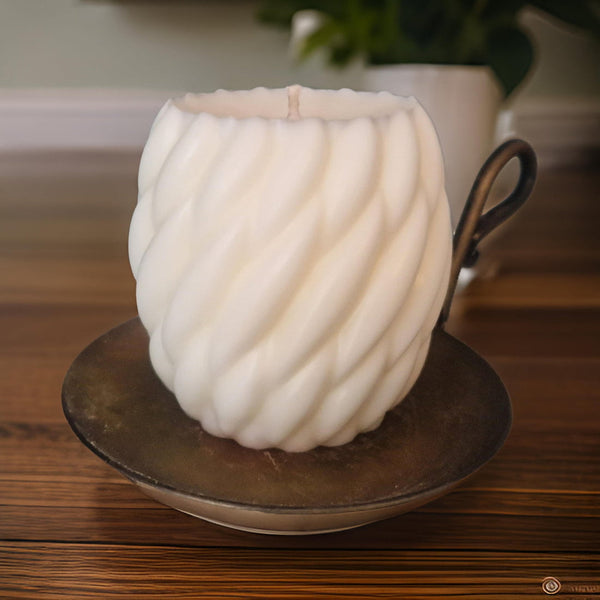 A white pillar candle with a spiral design is lit, sitting on a metal dish with a handle. The candle is indoors, providing a warm glow.