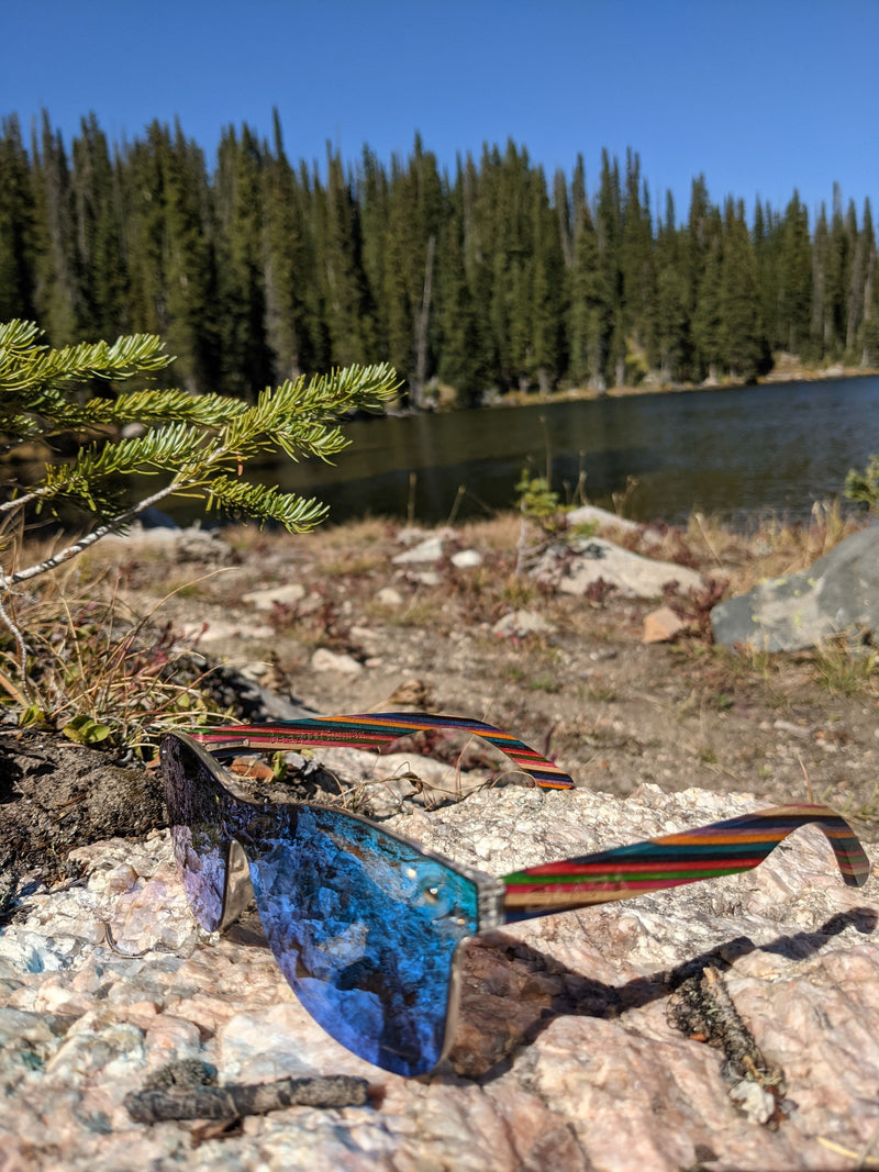 Emerald sky SAARA shades with rainbow recycled arms sitting on a rock in front of a lake