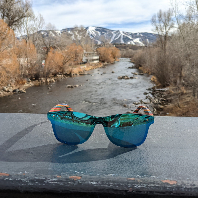 Emerald Sky SAARA shades sitting on a ledge over a river with the ski runs and mountains in the background