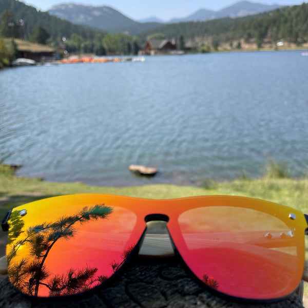 Bronco orange SAARA sunglasses with a reflection of evergreen branches in the shades, and a lake and mountains in the background. The glasses are orange and pink in color