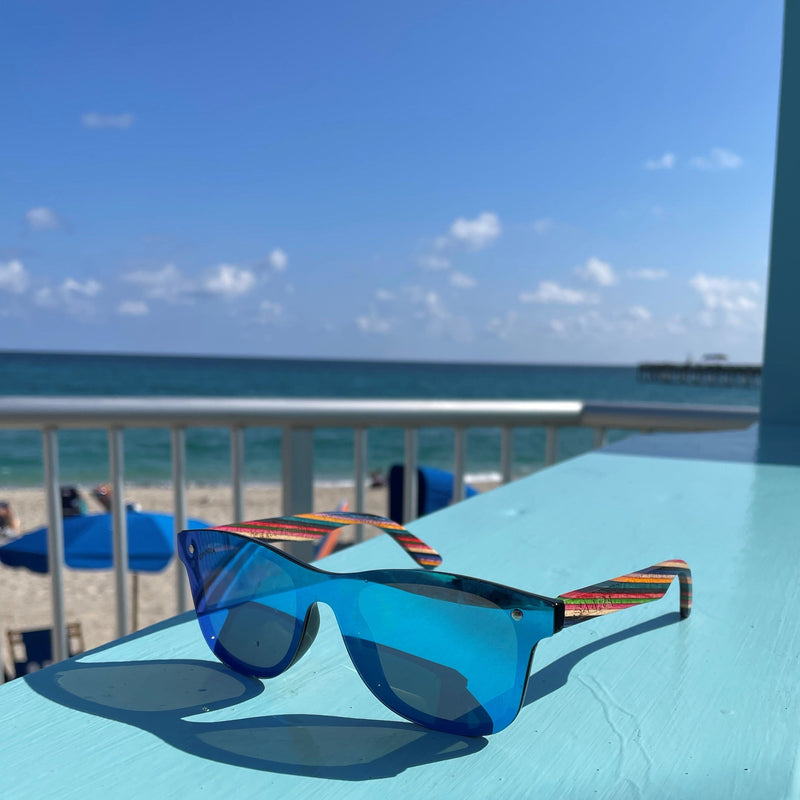 Blue SAARA shades with rainbow arms sitting on a blue ledge with the ocean in the background